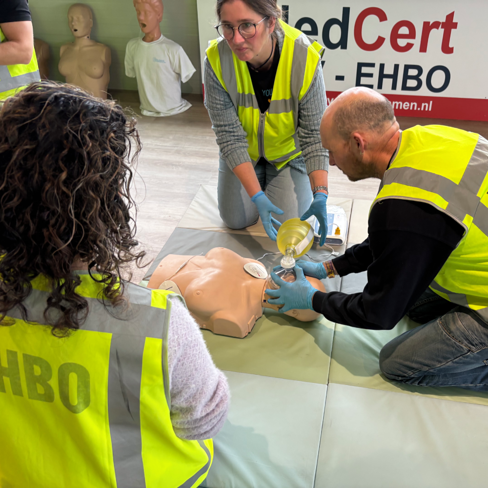 Twee cursisten oefenen ballonbeademing op een reanimatiepop tijdens een SEHSO-cursus van NedCert Experts in Emmen: de één houdt het masker met beide handen op het gezicht, de ander knijpt in de ballon, met een AED binnen handbereik.