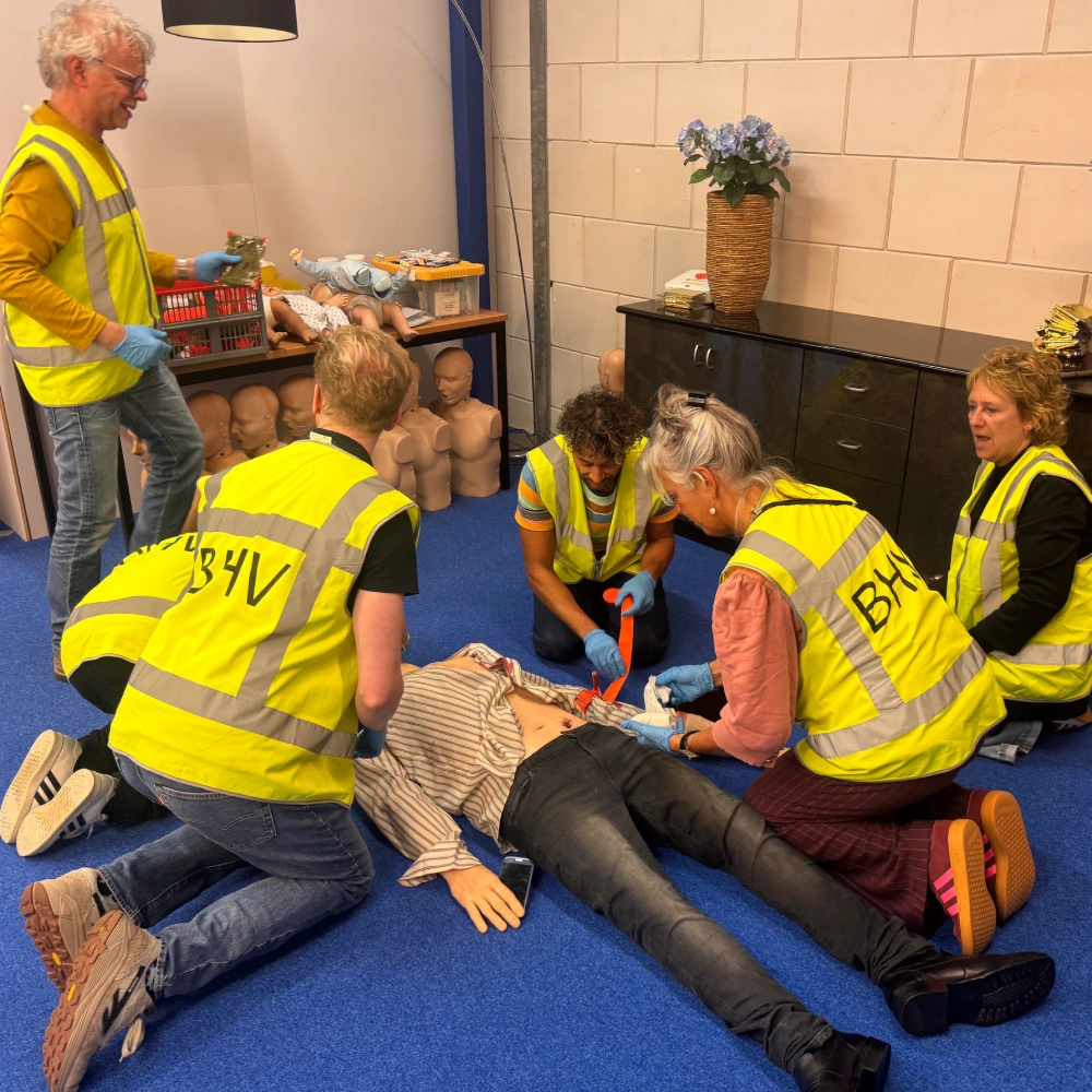 Cursisten in BHV-hesje verzorgen samen een slachtoffer dat op de grond ligt: de een drukt een wond af, een ander legt een tourniquet aan, terwijl de instructeur toekijkt.