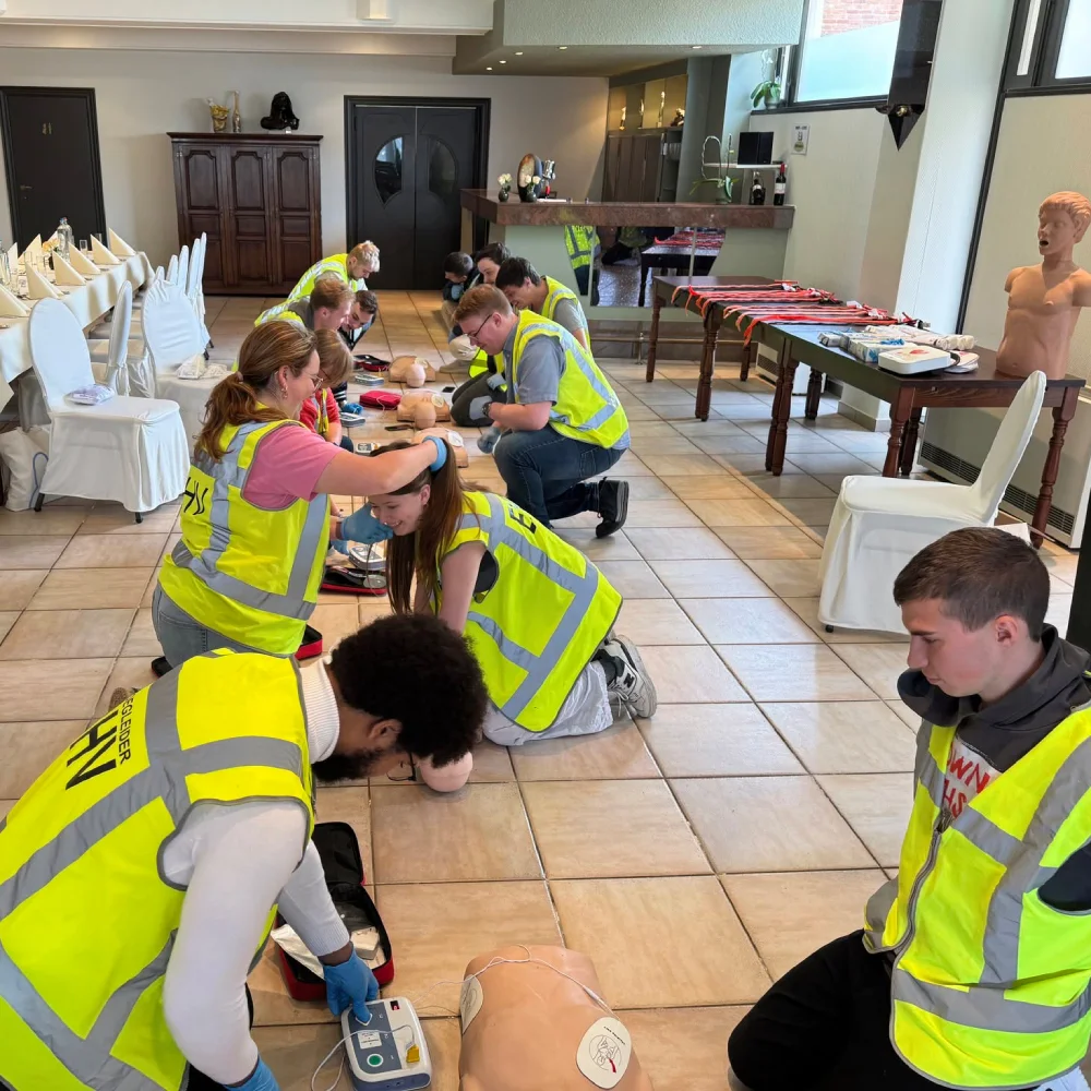 Cursisten in hesje oefenen in tweetallen reanimatie op oefenpoppen in de feestzaal van het eigen bedrijf: vooraan plakt een cursist de elektroden van een AED-trainer, ernaast controleert een ander de ademhaling met het hoofd van de pop gekanteld.