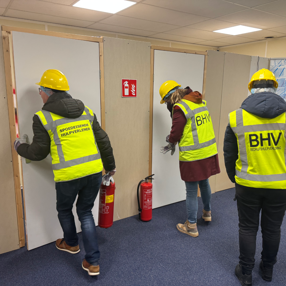 BHV'ers met helm en handschoenen voelen met de rug van de hand aan gesloten deuren tijdens een ontruimingsoefening, met een brandblusser binnen handbereik.