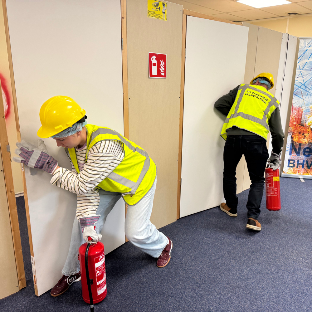 Twee cursisten met helm, handschoenen en BHV-hesje openen tijdens een ontruimingsoefening elk een deur, laag en vanaf de zijkant, met een brandblusser in de hand.