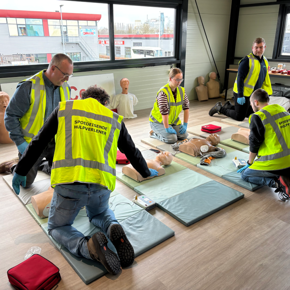 Cursisten in BHV-hesje oefenen reanimatie op oefenpoppen die op matten liggen, met een AED-trainer naast elke pop en een instructeur die meekijkt.