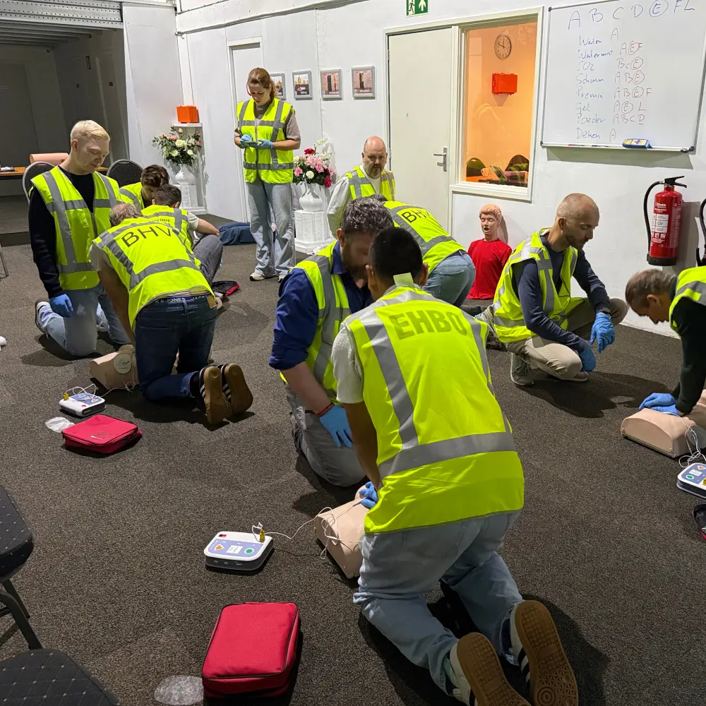 Een groep cursisten in BHV-hesje oefent tegelijk reanimatie op oefenpoppen met een AED-trainer ernaast, in een praktijkhal met een whiteboard over brandklassen en blusmiddelen aan de wand.