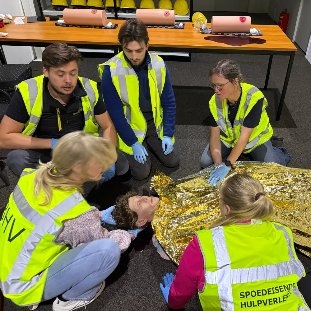 Vijf cursisten in BHV-hesje rond een oefenpop op de grond: een van hen houdt het hoofd stil, een ander dekt het lichaam af met een reddingsdeken, op de achtergrond staan wondtrainers op tafel.