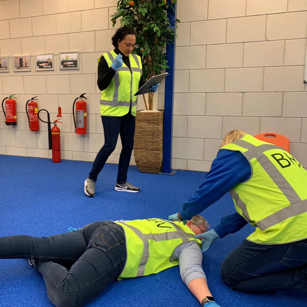 Een examinator met klembord beoordeelt een kandidaat in BHV-hesje die knielt bij een slachtoffer op de grond en het naar rugligging draait, met brandblussers aan de wand op de achtergrond.