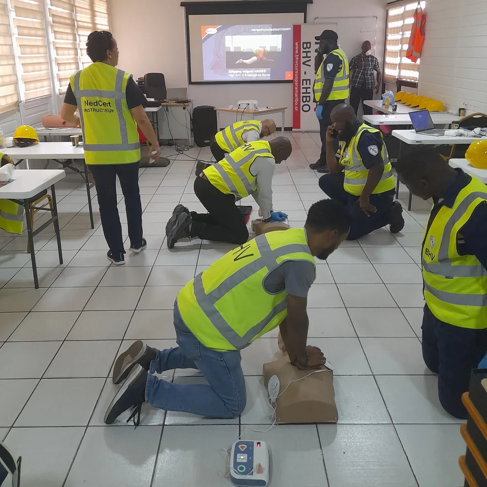 Cursisten in BHV-hesje oefenen reanimatie op oefenpoppen met een AED-trainer ernaast, terwijl een van hen met de telefoon aan het oor het alarmnummer belt en de instructeur meekijkt.