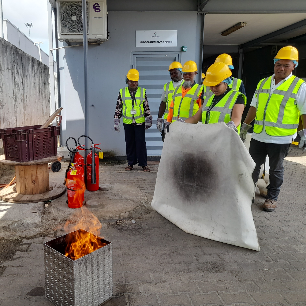 Cursisten leggen een blusdeken over een brandende oefenbak tijdens een incompany BHV-training op het terrein van een internationaal bedrijf.