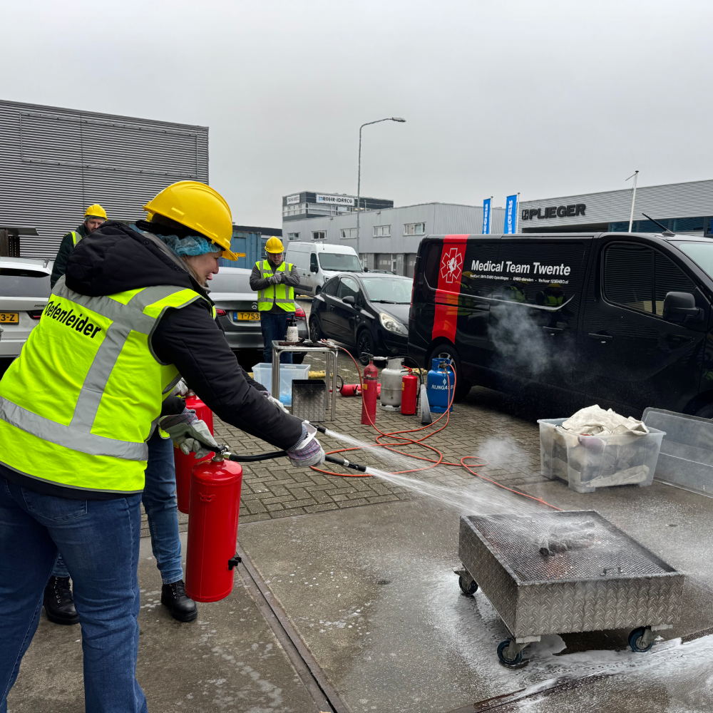 Cursist blust een oefenbrand met een schuimblusser op het terrein van een bedrijf, met blusmiddelen, gasfles en slang klaargezet op de achtergrond.