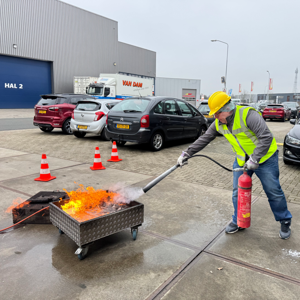Cursist blust een oefenbrand met een brandblusser op het parkeerterrein van een bedrijfspand op een industrieterrein.