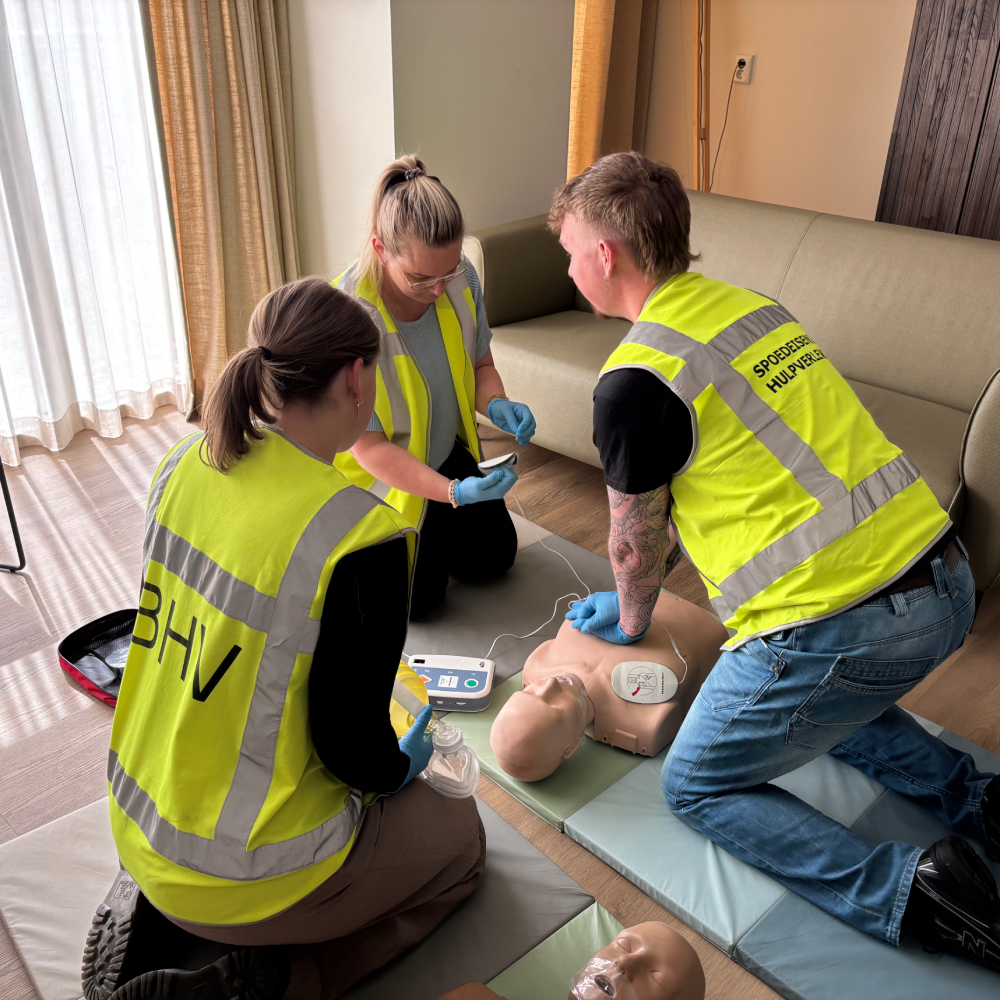 Drie cursisten reanimeren een oefenpop met AED en beademingsballon in de lounge van een bedrijfspand.