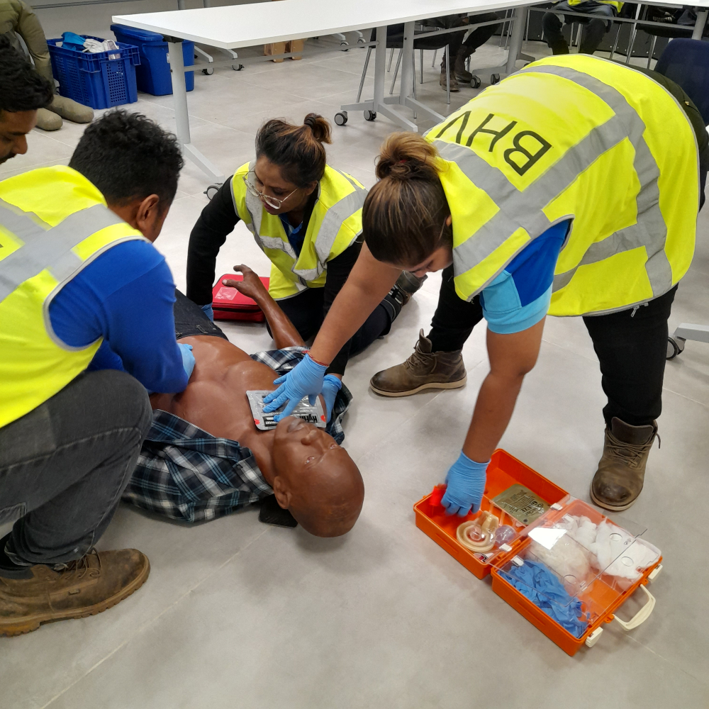 Meerdere cursisten verlenen samen hulp aan een oefenslachtoffer op de grond tijdens een BHV-cursus van NedCert Experts in Paramaribo, terwijl een van hen materiaal uit de EHBO-koffer pakt.