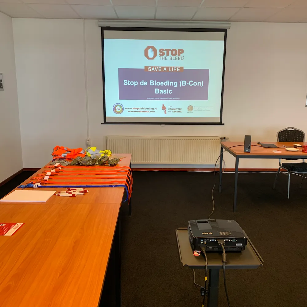 Cursusruimte klaargezet voor een Stop de Bloeding (B-Con) Basic training in 2016, met tourniquets en hesjes op tafel en de officiële cursuspresentatie van de American College of Surgeons op het projectiescherm.
