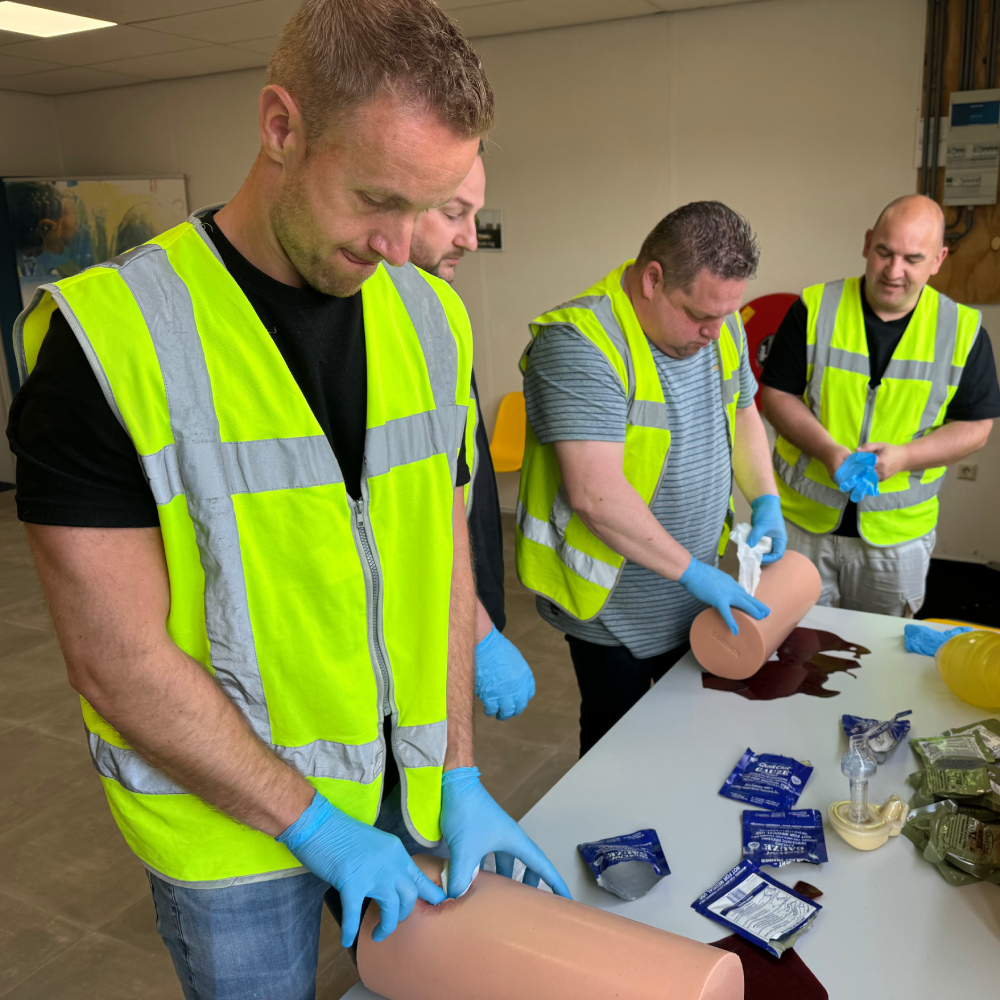 Cursisten in gele hesjes oefenen het opstoppen van een wond met hemostatisch gaas op oefenbenen tijdens een Stop de Bloeding training van NedCert Experts.